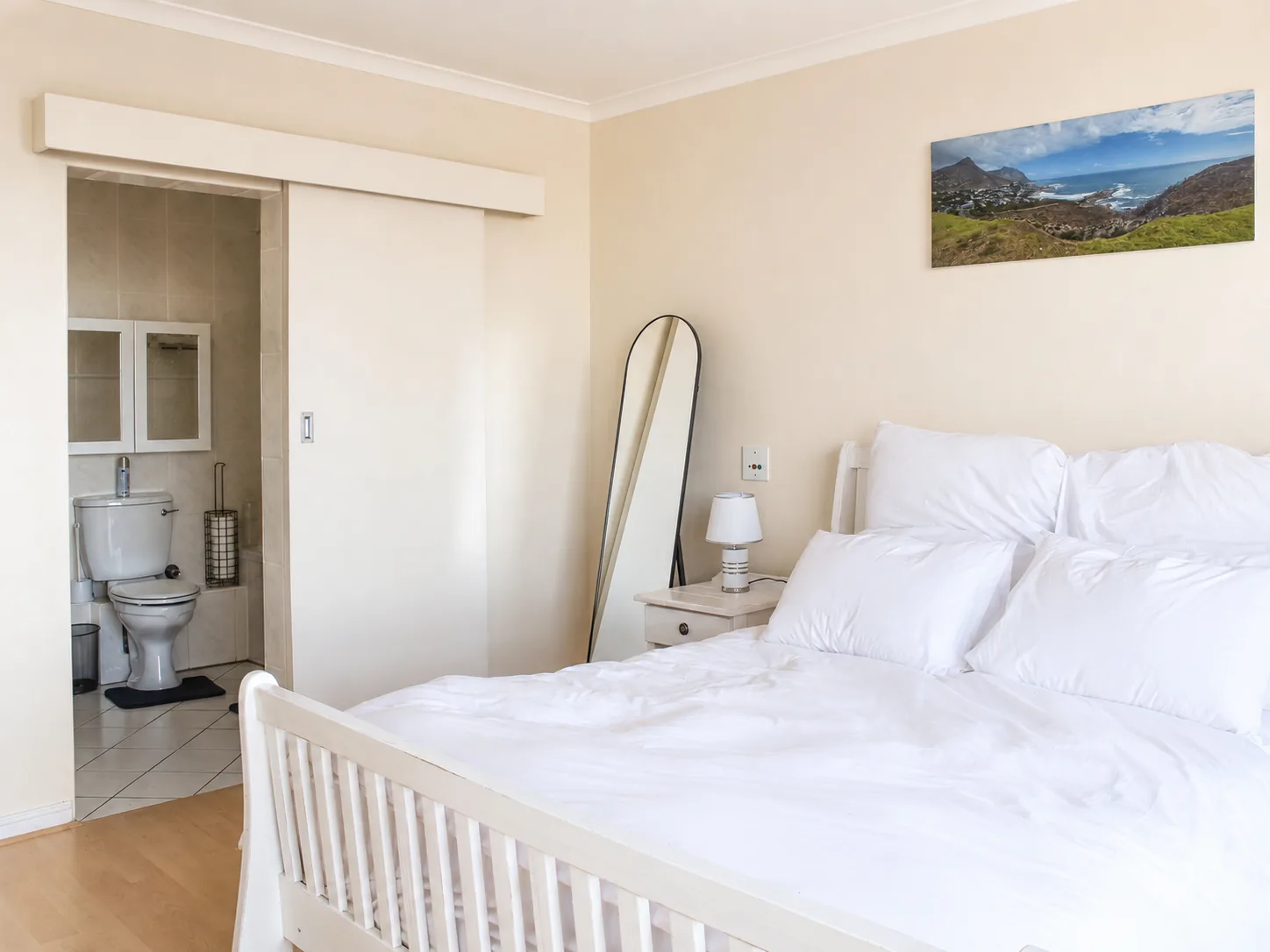 Bright bedroom with white bedding, full-length mirror and sliding door leading to an en-suite bathroom