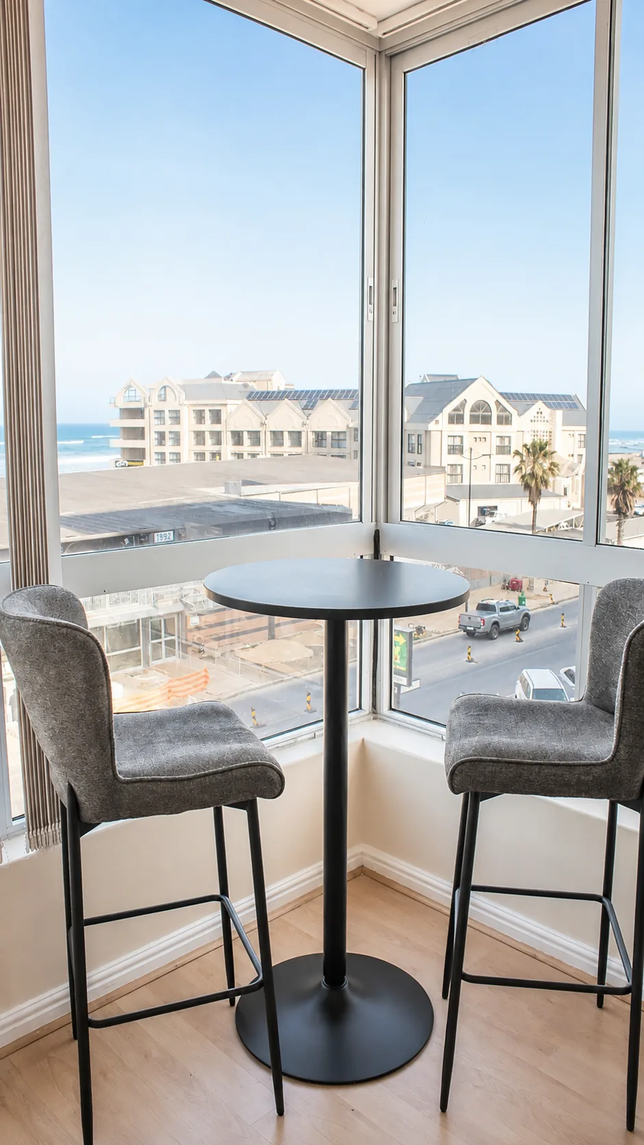 Two bar chairs and a round table beside corner windows with views of nearby buildings and the ocean
