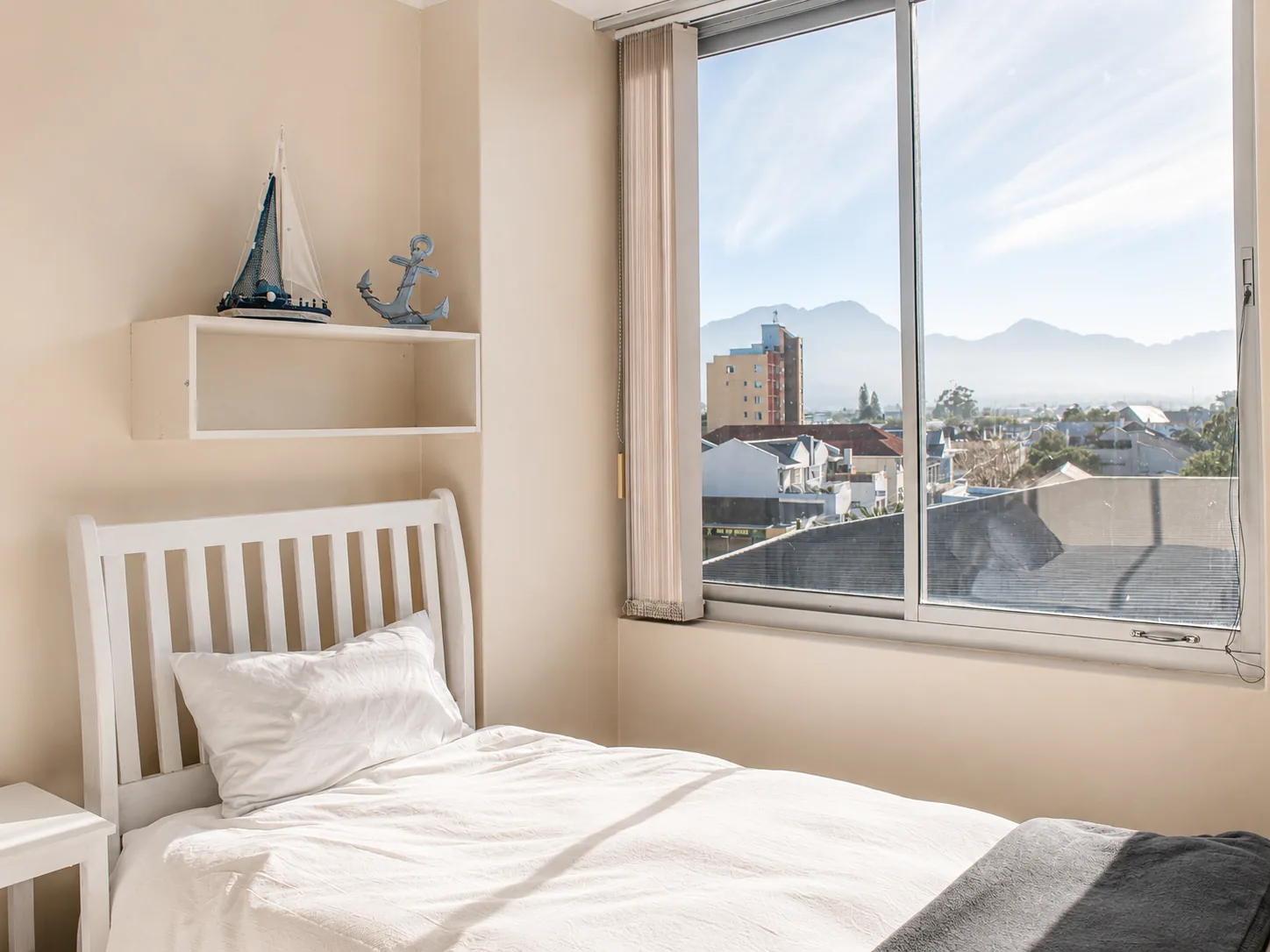 White single bed beside a large window overlooking nearby rooftops and a mountain range